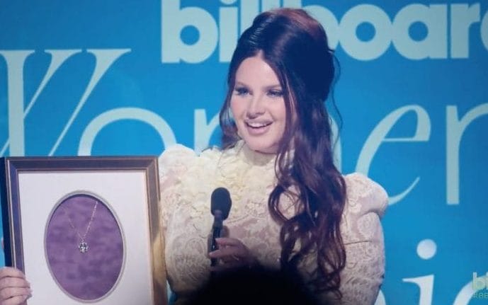 Lana Del Rey 2023 accepting Billboard's Women in Music Visionary award. Her hair is done up big and long, wearing a white dress.