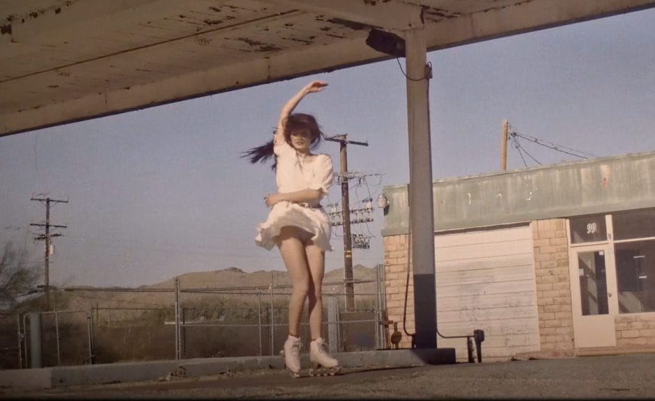Lana Del Rey rollerskaing in vacant gas station lot, wearing a white short dress. Hair covering face as she twirls in her skates. her arm is up to the sky.