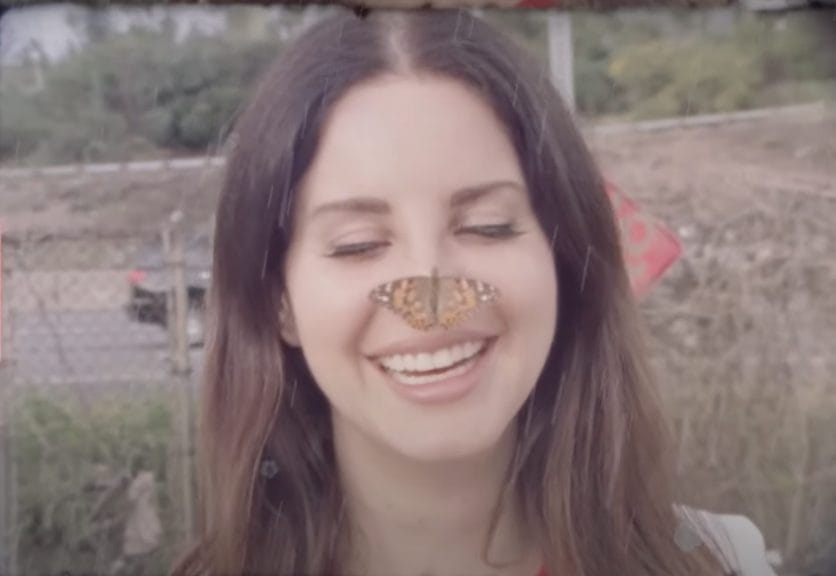 Close Up, Lana Del Rey has a small live butterfly on her nose, she is smiling and eyes closed.