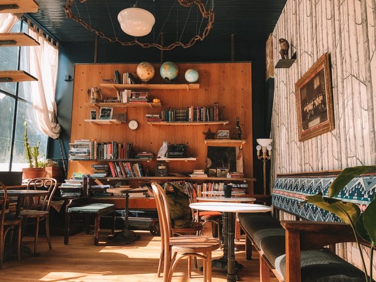 Image shows a coffee shop that contains plants, couches, and a wall filled with books. You can see a coffee cup sitting on one of the tables.