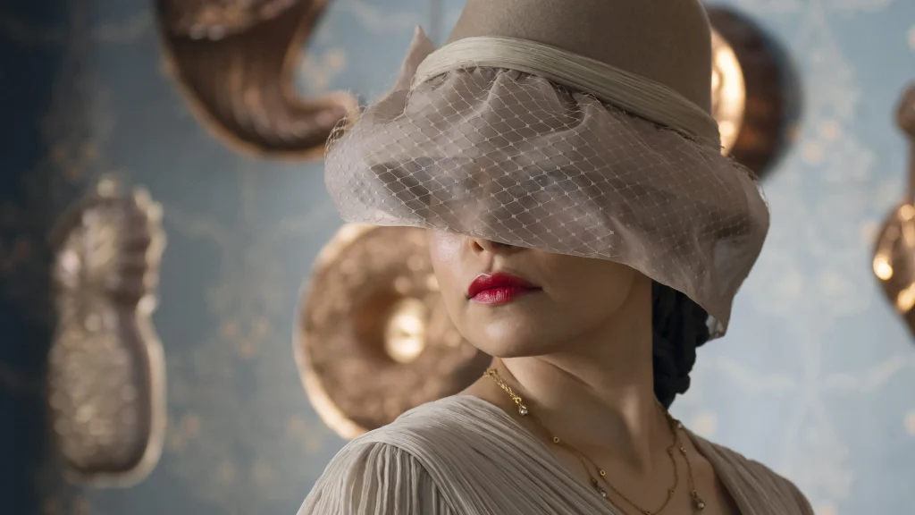 Jessica Parker Kennedy as Medusa, wearing a cream coloured dress and a hat that purposefully obscures her eyes. 