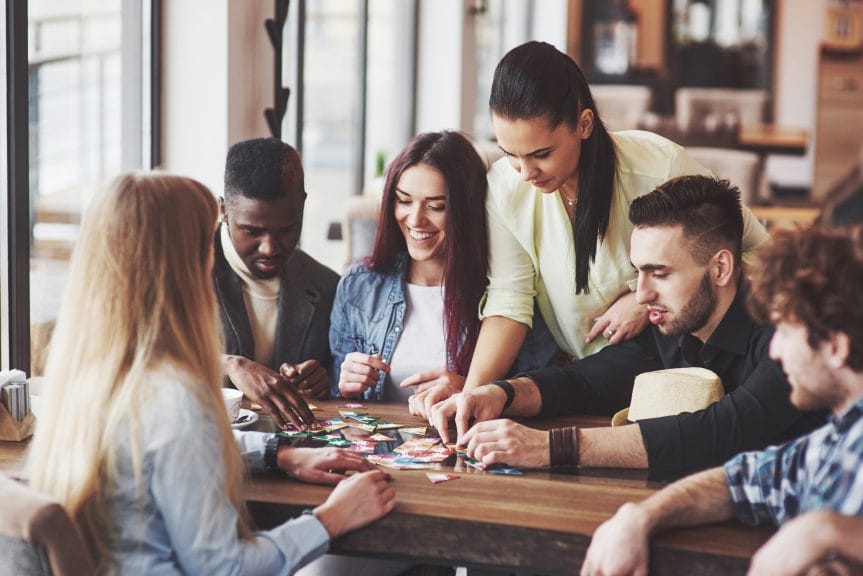 A group of young men and woman, around a table, playing a board game. The social and mental aspect of games can be a huge help to your mental health and wellness.