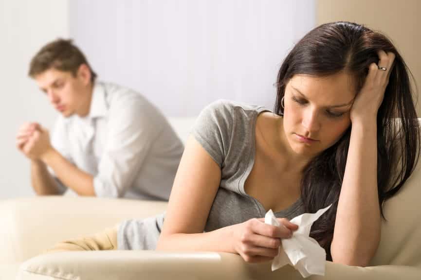 A young couple sit opposite each other on the couch. The woman is closer to the camera, holding a tissue with her head in her hands.