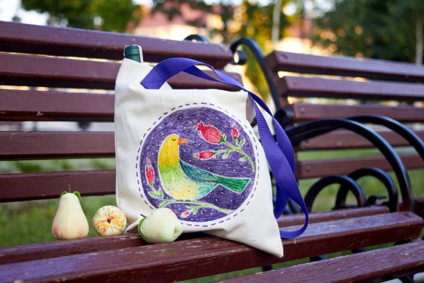 Spring Tote Bag hand painted with a green and yellow bird and red flowers on a purple background. Sitting on an outdoor bench.