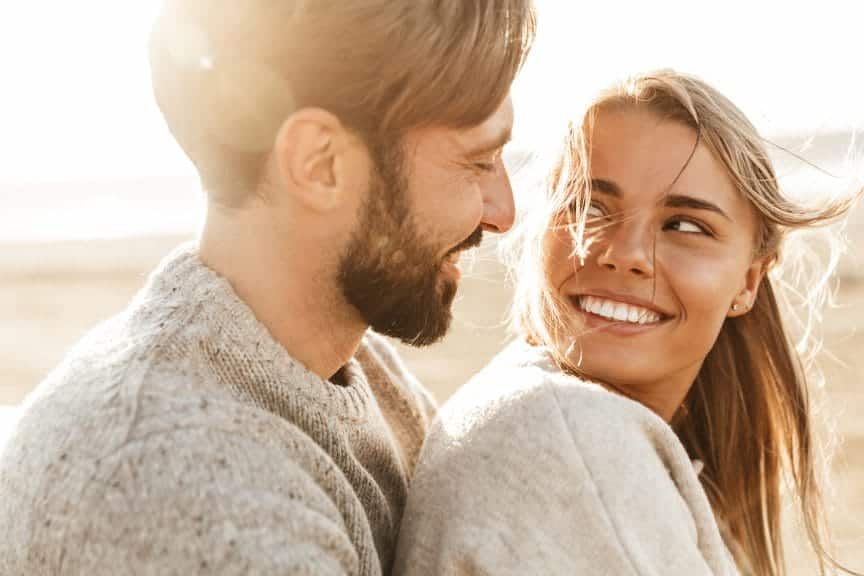 A young couple are looking at each other, smiling. The sun is setting behind them.