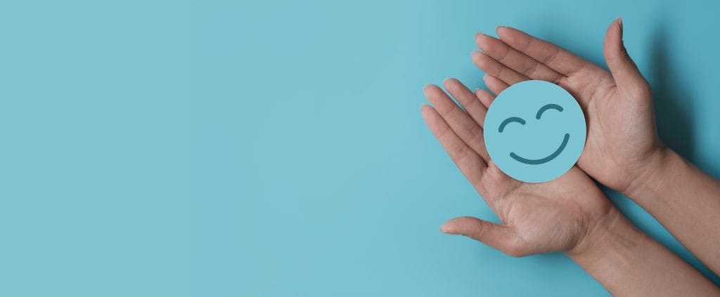 A baby blue background, with two hands holding a blue circle smiling paper face. 