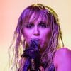 A woman with dark eyeshadow and wet looking blonde hair stands in front of a yellow background holding a microphone with a black leather glove