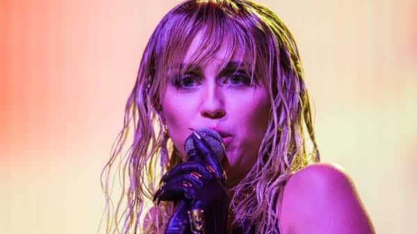 A woman with dark eyeshadow and wet looking blonde hair stands in front of a yellow background holding a microphone with a black leather glove