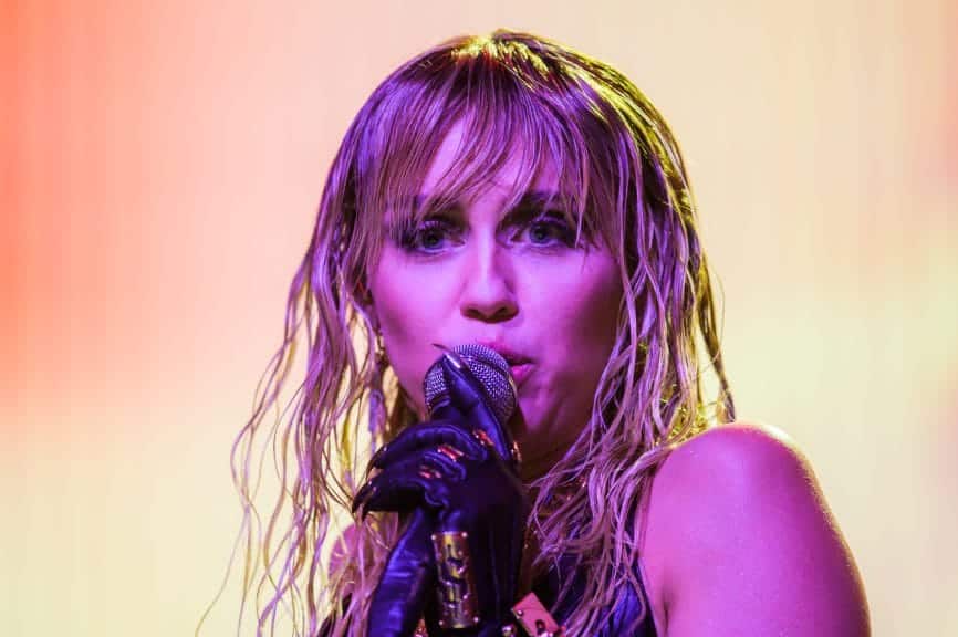 A woman with dark eyeshadow and wet looking blonde hair stands in front of a yellow background holding a microphone with a black leather glove