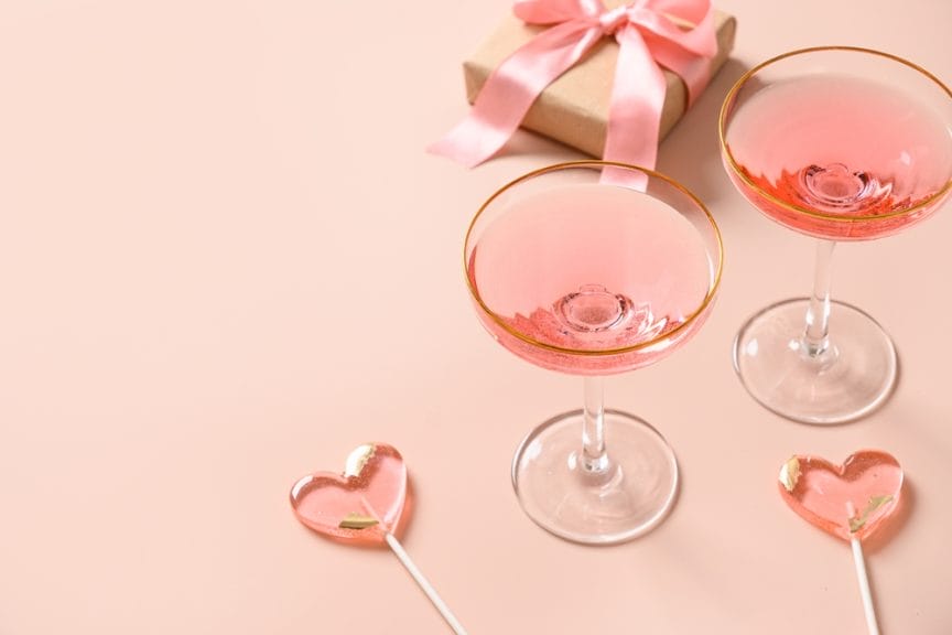 Festive champagne, romantic gift and two pink lollipops on stick on pink background. Galentine's day greeting card. Close up.