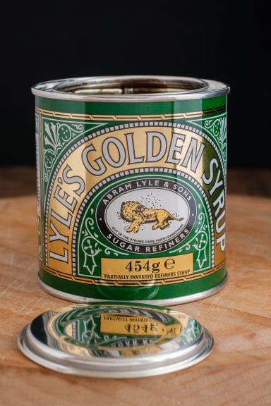 a green and gold tin sits on a wooden worktop. It reads 'Lyle's Golden Syrup' and features an illustrated dead lion surrounded by honeybees