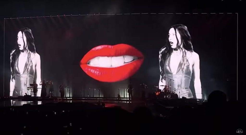 Olivia Rodrigo onstage with two images of her on screen and red lips image in the center.