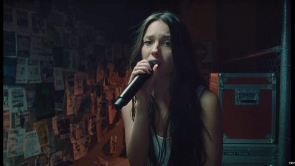 Olivia Rodrigo sings into a microphone within a darkened room.