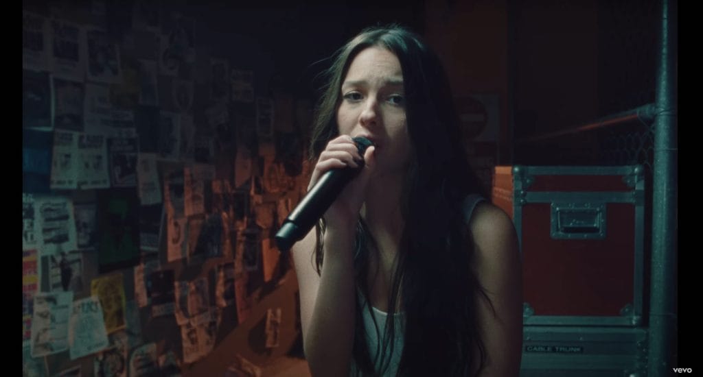 Olivia Rodrigo sings into a microphone within a darkened room.