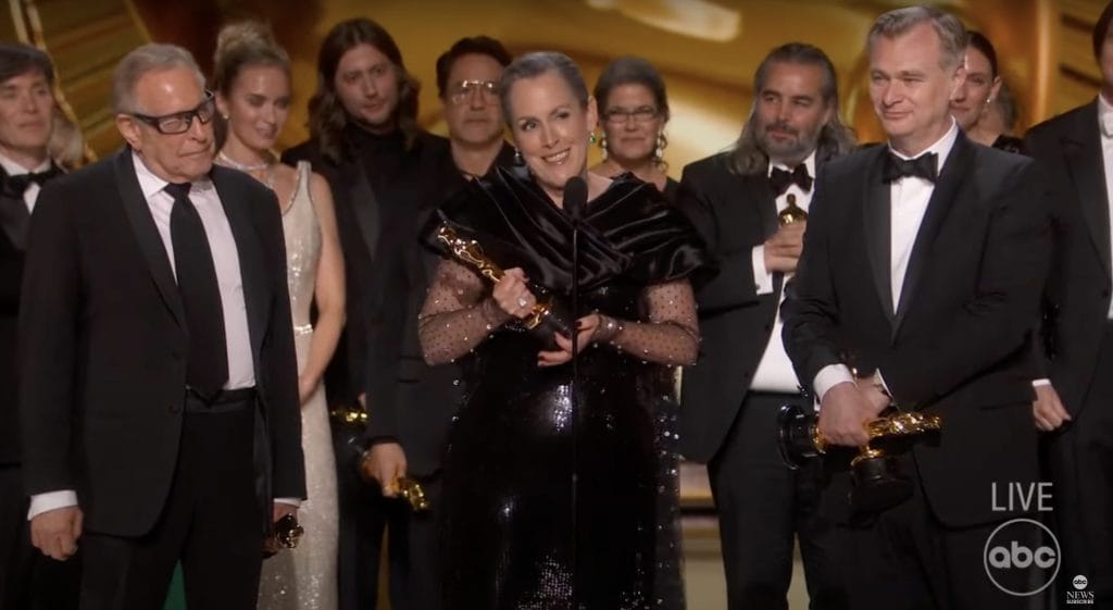Eleven 'Oppenheimer' cast members onstage in formal attire. Woman in a black dress stands in front of the microphone holding an Oscar trophy. 