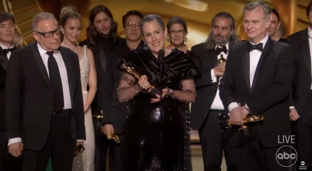 Eleven 'Oppenheimer' cast members onstage in formal attire. Woman in a black dress stands in front of the microphone holding an Oscar trophy.
