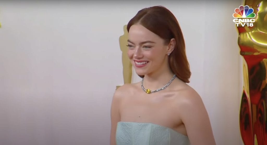 Emma Stone smiles in a light blue dress with a silver necklace on the Oscars red carpet. 