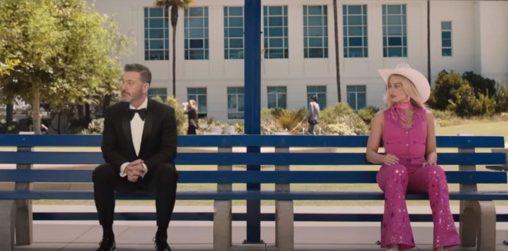 Jimmy Kimmel sits on a bench next to Margot Robbie acting as "Barbie" from the movie Barbie.