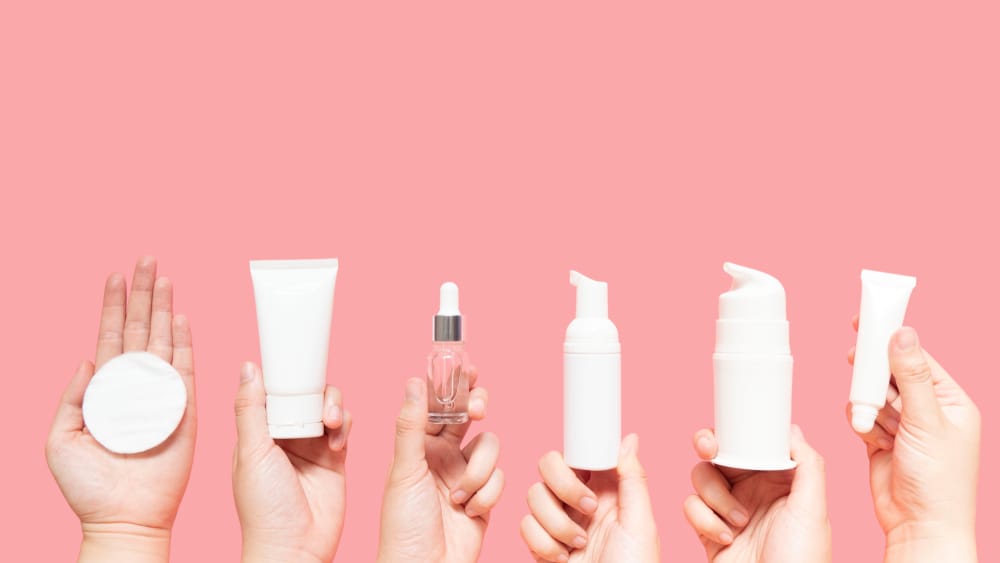 Hands holding different skin care products against a pink backdrop