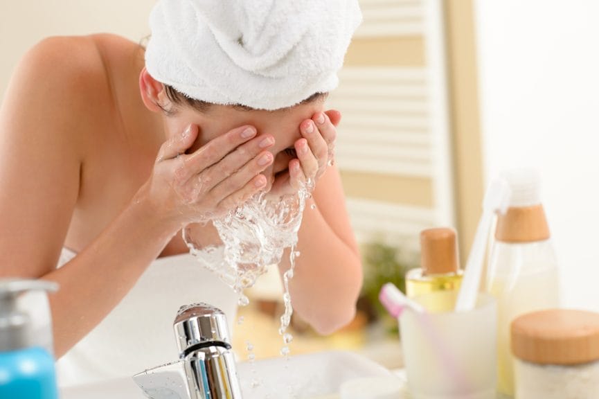 A woman bending over a sink washing skin care products off her face with water in a bathroom