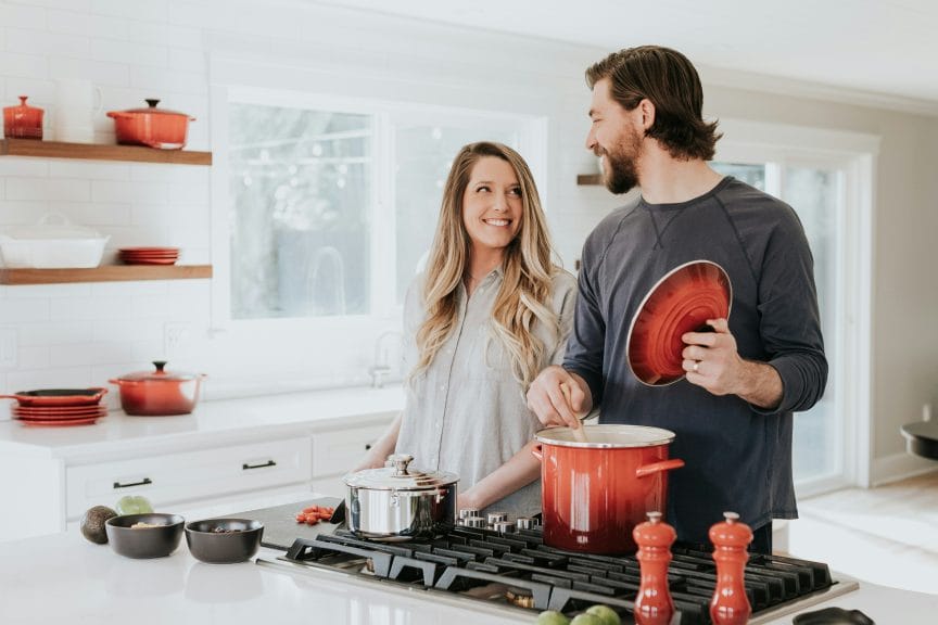 man and woman making dinner in red pots at the stove. They're looking at each other and smiling
