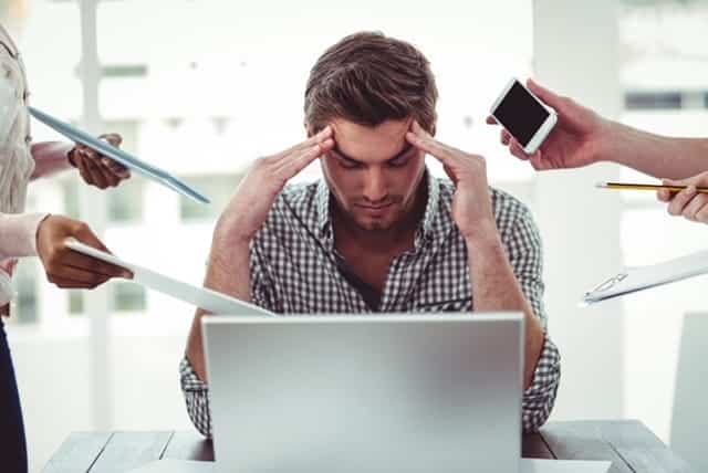 A man with his hand to his temples and cellphone by his ear, stressfully staring at his laptop.