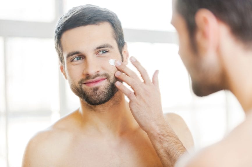 Handsome man applying moisturizer cream to his face in the mirror