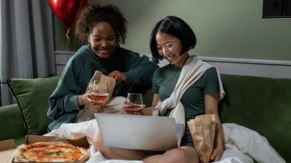 Two women are sitting on a couch watching something on a laptop.