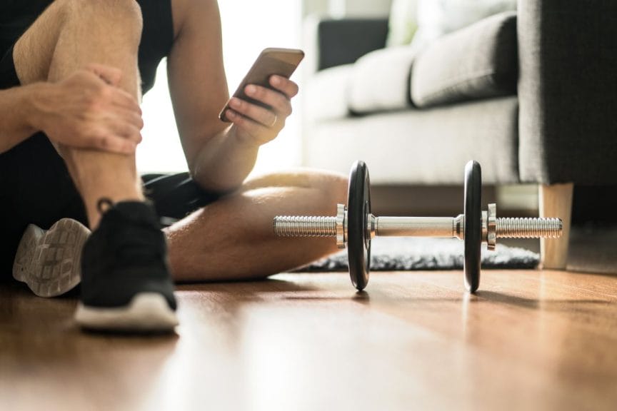 A man sitting on his living room floor looking at his phone with weights sitting beside him.