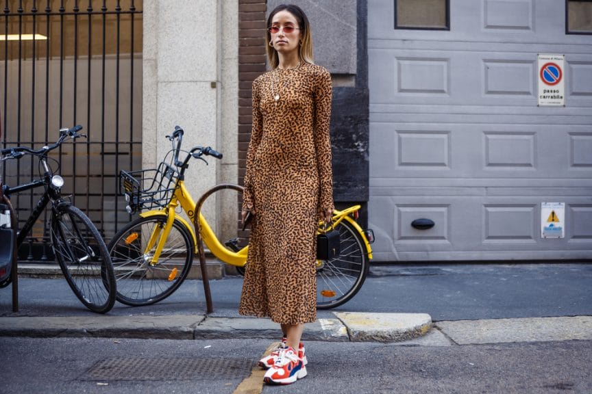 The picture shows a woman in an animal printed long dress. She is standing in the street and there is a yellow bike behind her. She is wearing a pair of sunglasses.