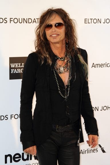 Steven Tyler standing in front of a white wall with text behind it.