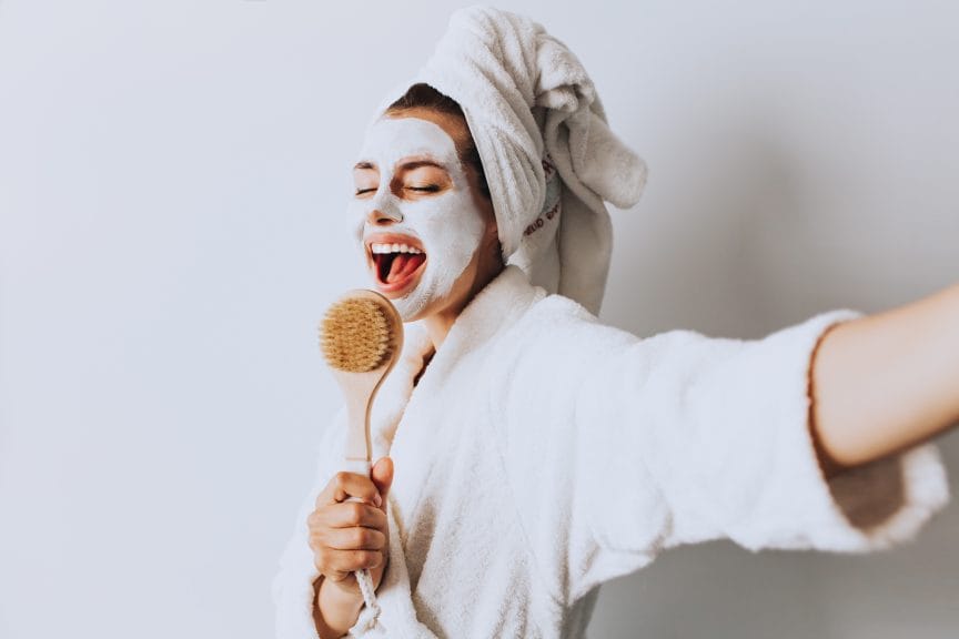 A girl in a bathrobe, hair up in a towel, and wearing a facemask holds up a brush like she's singing into it.