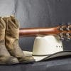 Cowboy boots with a guitar placed on top of a cowboy hat set on the floor against a grey background