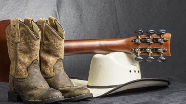 Cowboy boots with a guitar placed on top of a cowboy hat set on the floor against a grey background