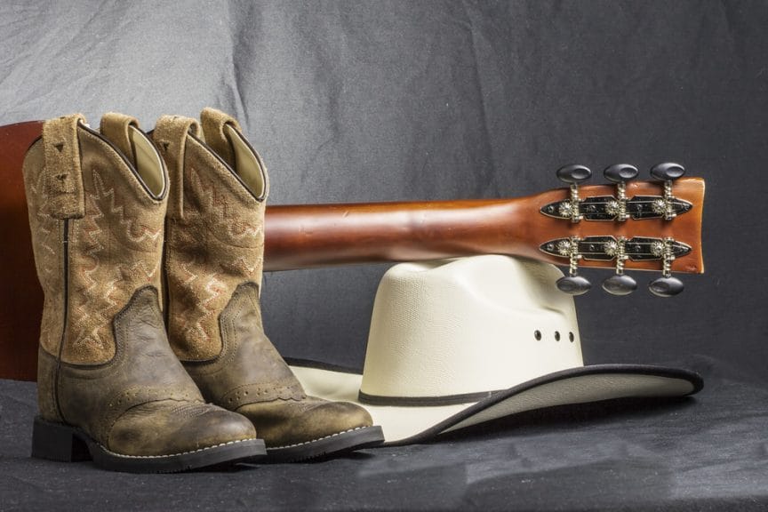 Cowboy boots with a guitar placed on top of a cowboy hat set on the floor against a grey background