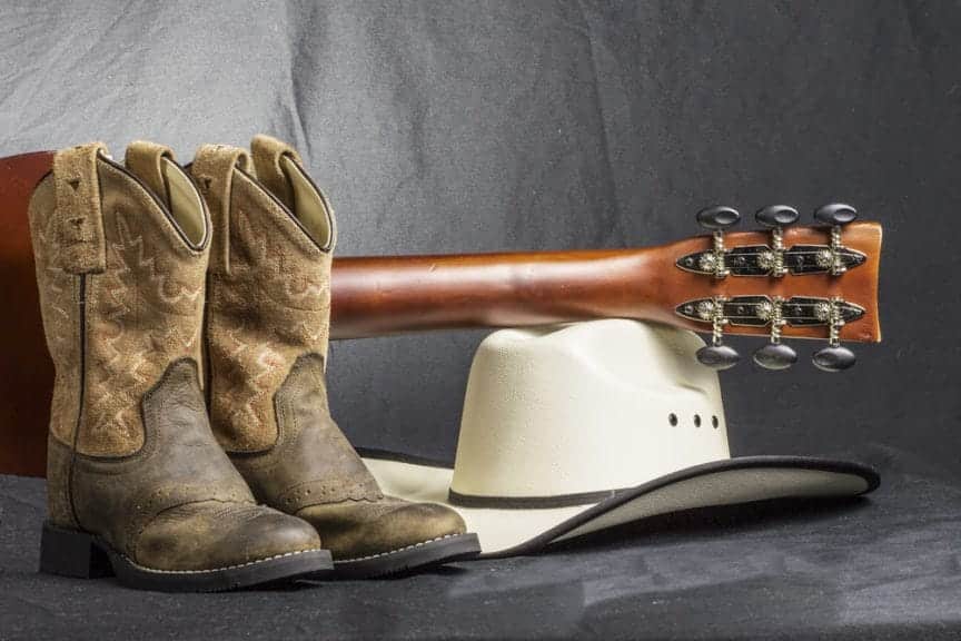 Cowboy boots with a guitar placed on top of a cowboy hat set on the floor against a grey background