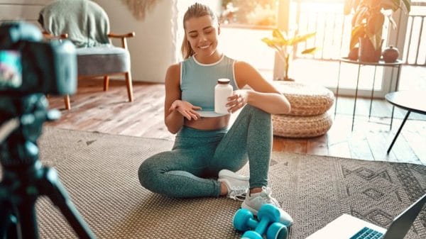 A blogger sitting on the floor with leg bells and a laptop in front of her showing a sports bottle to the camera in a living room.