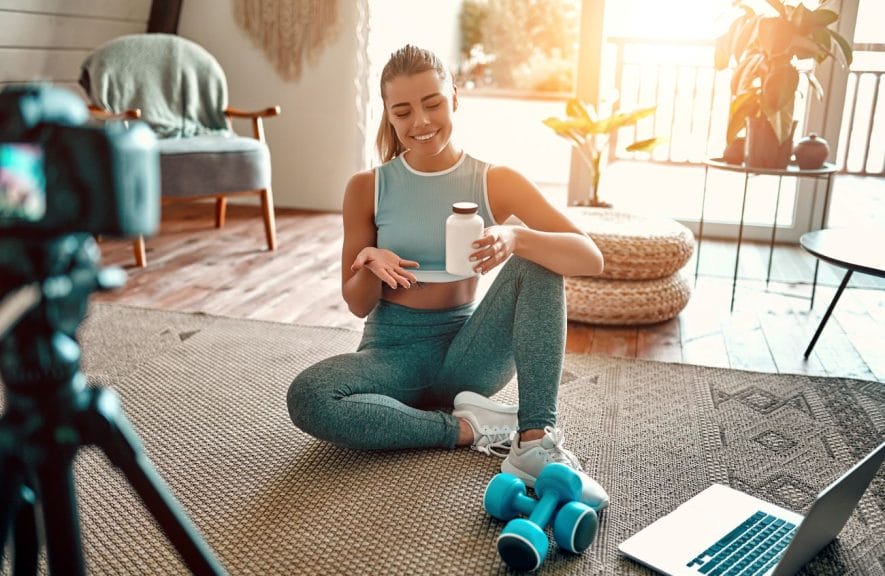 A blogger sitting on the floor with leg bells and a laptop in front of her showing a sports bottle to the camera in a living room.