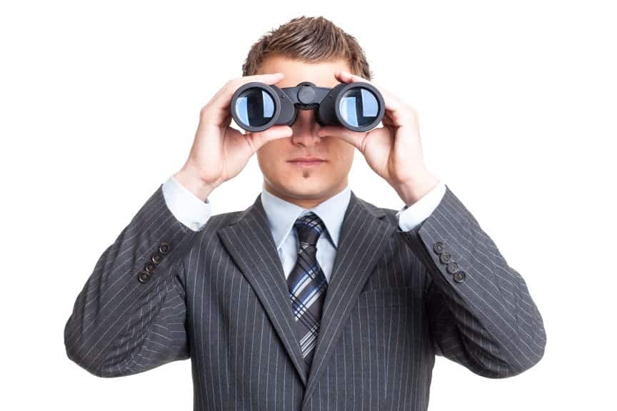 A young man in a gray suit with a pair of binoculars being held up to his eyes. 