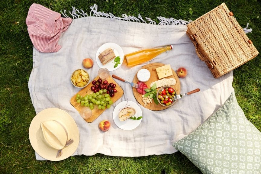 Food on a white picnic blanket with a pillow, hat and a picnic basket.