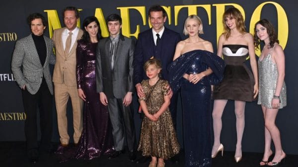 Actor Bradley Cooper stands with the cast of Maestro in front a poster that says Maestro in yellow lettering. His daughter is stood in front of him, she is seven years old, blonde and in a long leopard print dress