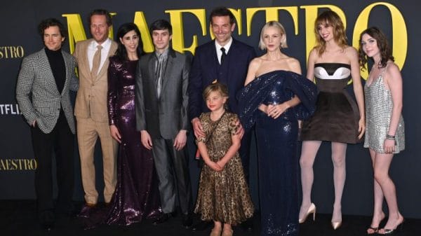 Actor Bradley Cooper stands with the cast of Maestro in front a poster that says Maestro in yellow lettering. His daughter is stood in front of him, she is seven years old, blonde and in a long leopard print dress
