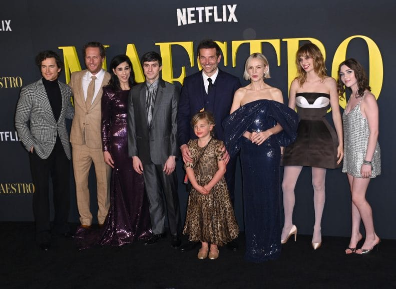 Actor Bradley Cooper stands with the cast of Maestro in front a poster that says Maestro in yellow lettering. His daughter is stood in front of him, she is seven years old, blonde and in a long leopard print dress