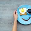 breakfast on a blue plate that consists of toast with a heart-shaped fried egg, a cup of coffee, and a line of sauce that forms a smiley face, Hands are placed gently on the sides of the plate