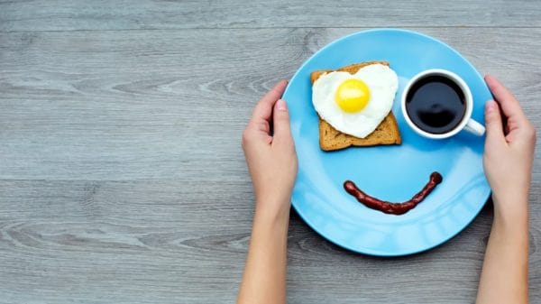 breakfast on a blue plate that consists of toast with a heart-shaped fried egg, a cup of coffee, and a line of sauce that forms a smiley face, Hands are placed gently on the sides of the plate