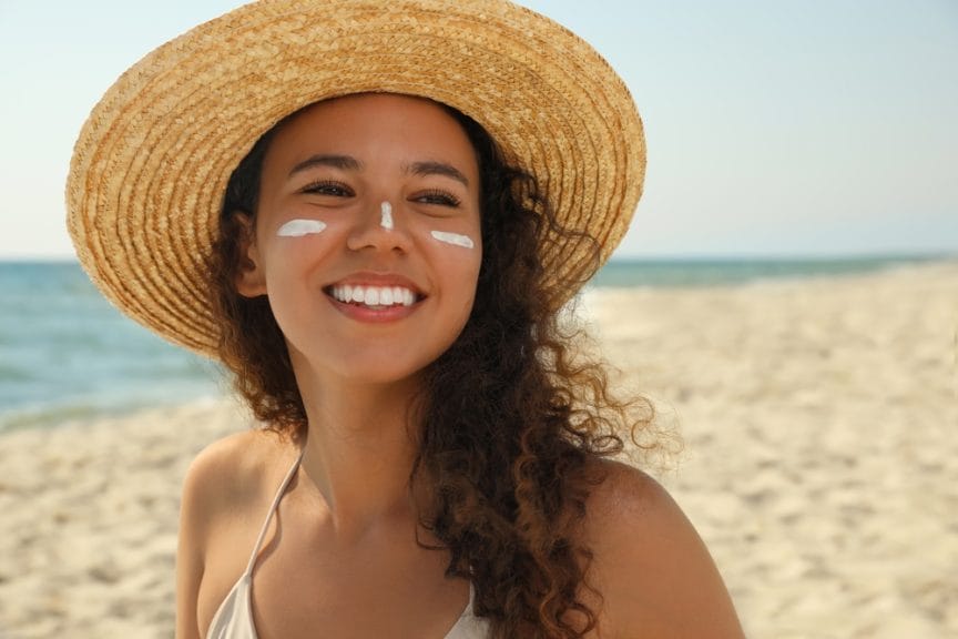 woman on the beach wearing sunscreen on her nose and cheeks smiling with a hat