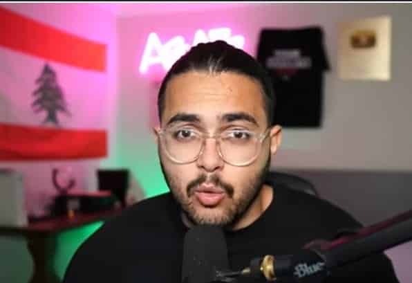 An Arabian man with black hair and glasses sits in front of a microphone wearing a black t-shirt in front of a pink and green neon background