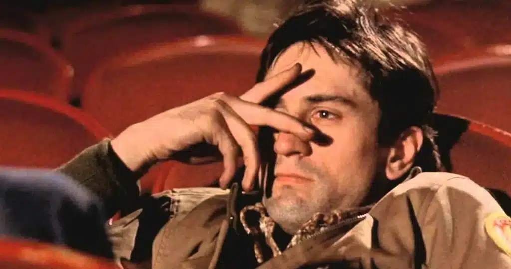 Robert De Niro, as Travis Bickle, hikes half his face while stitting in a red-seated movie theater.