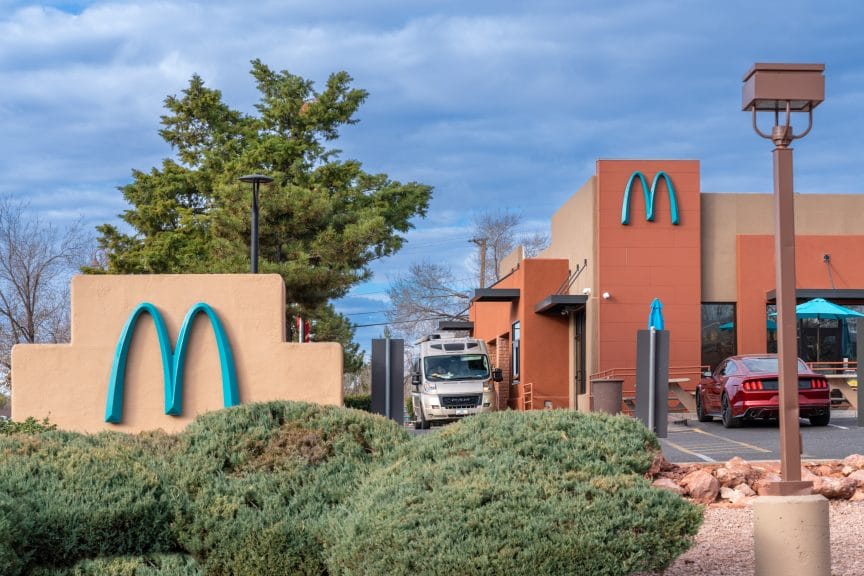 Pale red buildings in front of a blue sky. There is a large blue M on both buildings with a tree behind them and some shrubs in front of them.