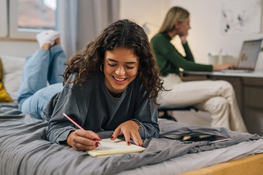 High school students that became college roommates doing homework in their dorm while smiling.